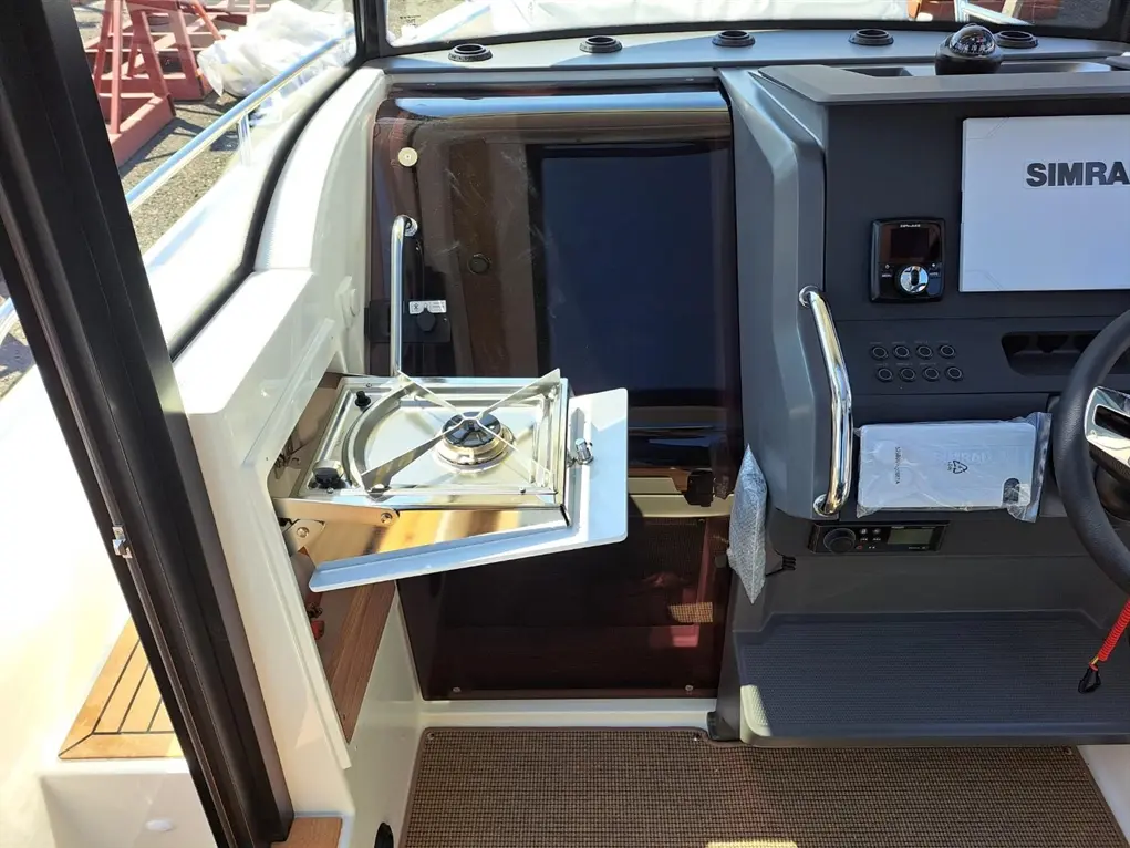 Interior of a boat showing a mounted SIMRAD display, a stove, and control panel with buttons and a fabric-wrapped steering wheel.